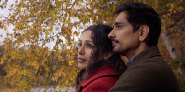 "A couple stands close together outdoors, looking thoughtfully into the distance surrounded by autumn trees with yellow leaves, suggesting a peaceful or contemplative moment in a natural setting."